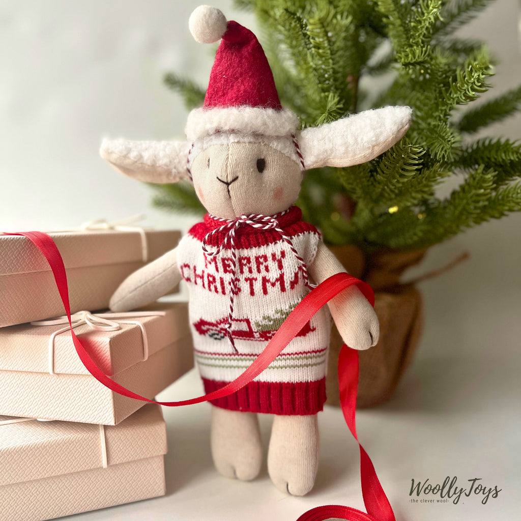 Kézműves levendula illatú bárány baba áll egy karácsonyfa előtt, mellette ajándékdobozok, kezében egy piros szalaggal, ahogy ajándékot csomagol. A bárányon egy piros fehér színű, autó mintás kötött mellény és egy piros mikulás sapka van. 