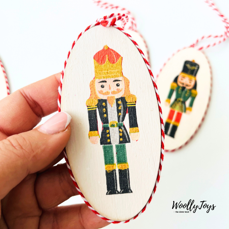 Egy kéz tartja a kézműves diótörő mintás fa karácsonyfadíszt, közel fókuszban, bemutatva a dísz méretét és finom kidolgozását.