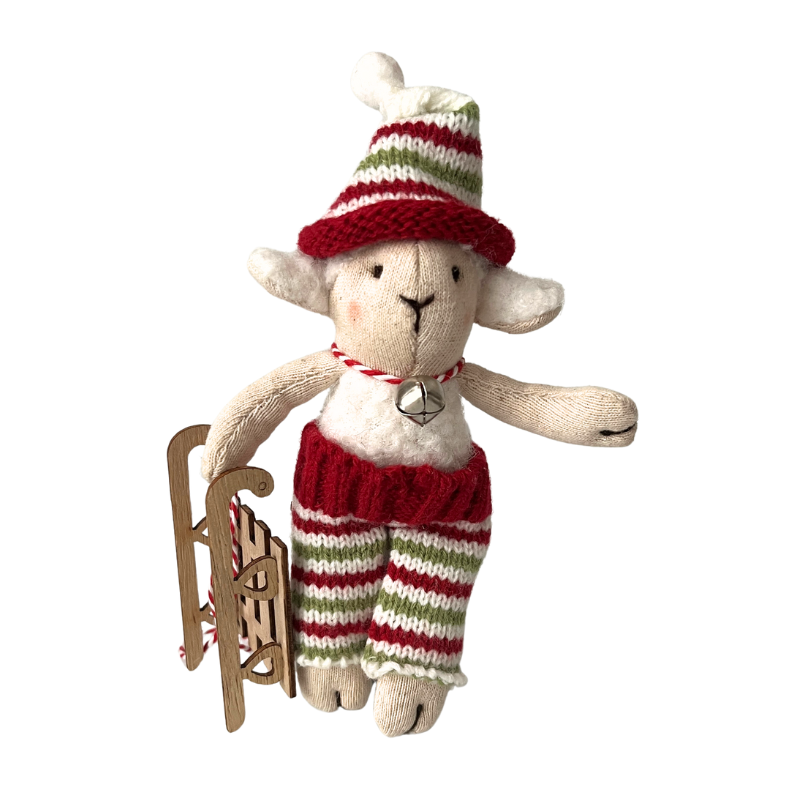 Öltöztethető kézműves fehér szőrméjű bárány figura piros fehér zöld csíkos kötött nadrágban és csíkos sapkában. A bárány egy kis fa szánkót tart a kezében, nyakában egy fém csengettyű.