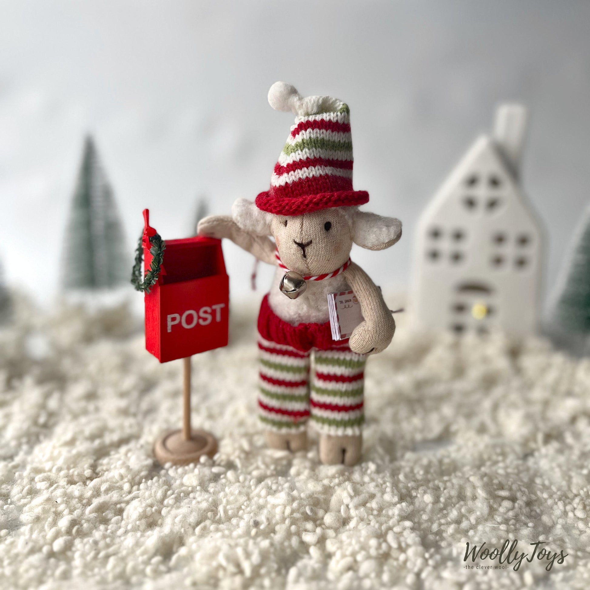 Felhőcske fehér színű mozhatható bárány figura a kezében tartja a leveleket, amiket épp készül bedobni  a kézműves piros fa Mikulás állványos postaládába. A táj havas, téli, a háttérben egy fehér ház és zöld fenyők sziluettje látszik. 