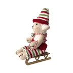 Gyapjúval töltött fehér bárány figura karácsonyi kötött ruhaszettben egy fa szánkón ül.  Kötött csíkos nadrágban és kötött sapkában. Nyakában fém csengettyű. 