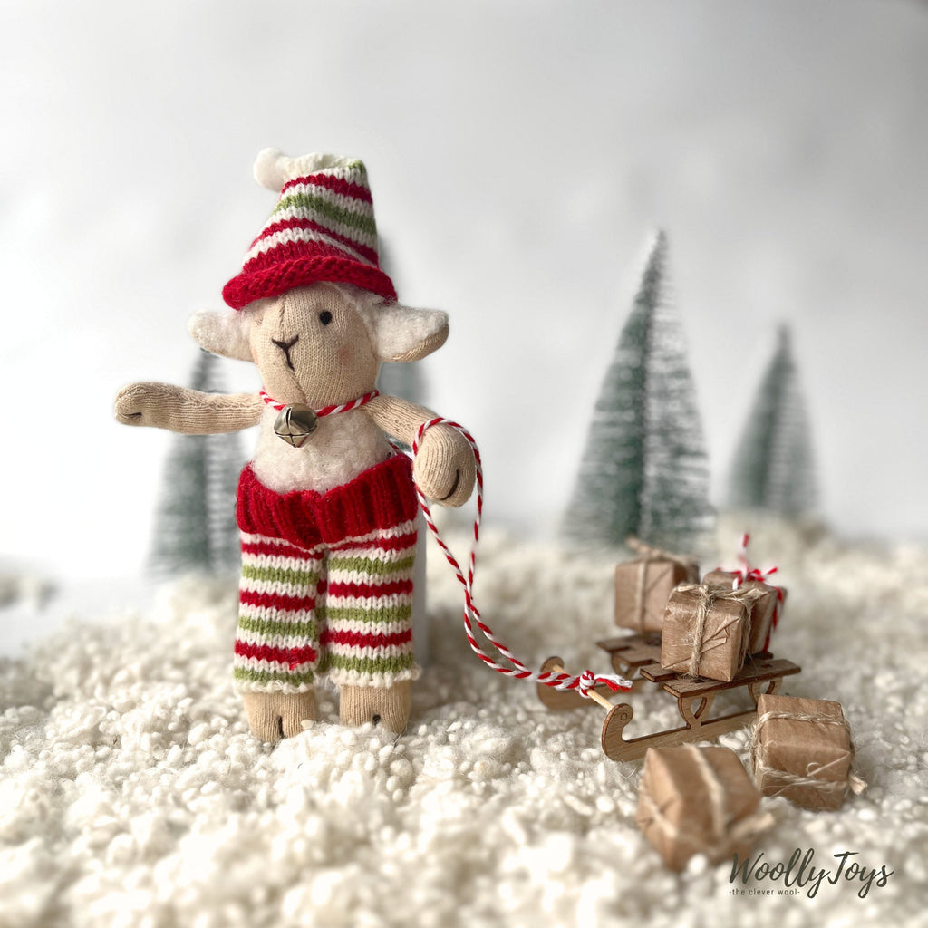 Kézműves fehér színű bárány figura, lába és karja mozgatható, csíkos kötött karácsonyi nadrágban és sapkában fa szánkót húz, mely tele van ajándékokkal. A képen havas a táj, hátterében zöld fenyők állnak. 