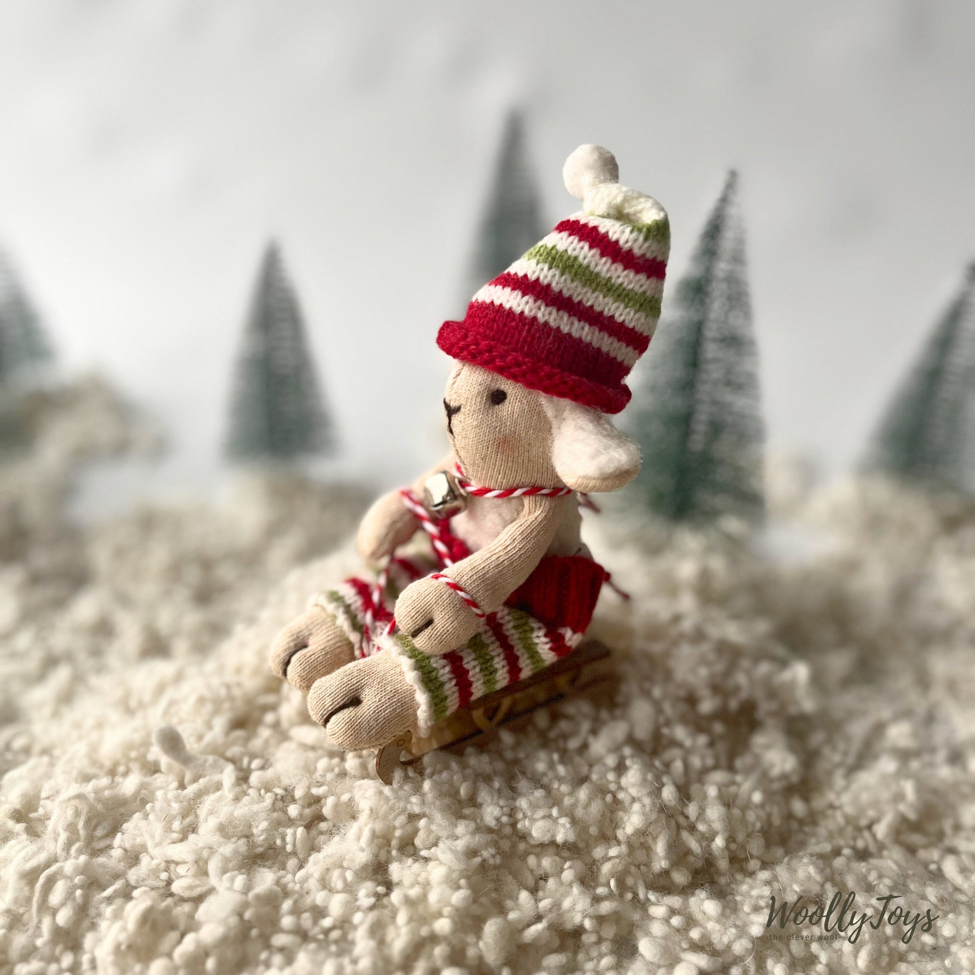 Felhőcske fehér színű bárány figura karácsonyi kötött nadrágjában és sapkájában fa szánkóján ül. Nyakában fém csengettyű. A táj téli hangulatú,  havas, hátterében zöld fenyők állnak. 