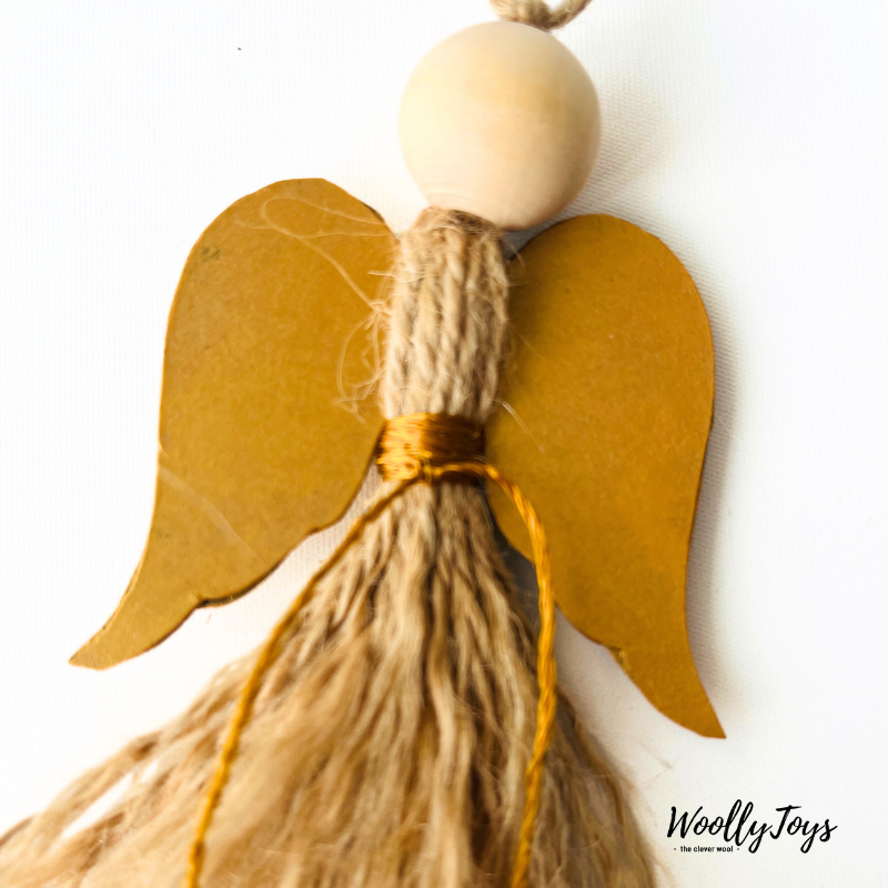 Közelkép egy kézműves natúr angyal karácsonyfadíszről arany szárnnyal, kiemelve a spárga textúráját és a fa golyó fej natúr jellegét.