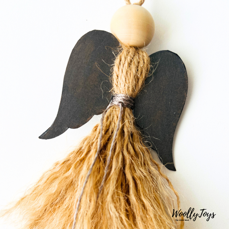 Közelkép egy kézműves natúr angyal karácsonyfadíszről ezüst szárnnyal, kiemelve a spárga textúráját és a fa golyó fej natúr jellegét.