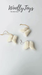 Videó, amelyen egy kéz mutatja be a fehér kézműves gyapjú  angyalszárny karácsonyfadísz minden oldalát, kiemelve a finom textúrát és a gravírozható fa hátoldalt.